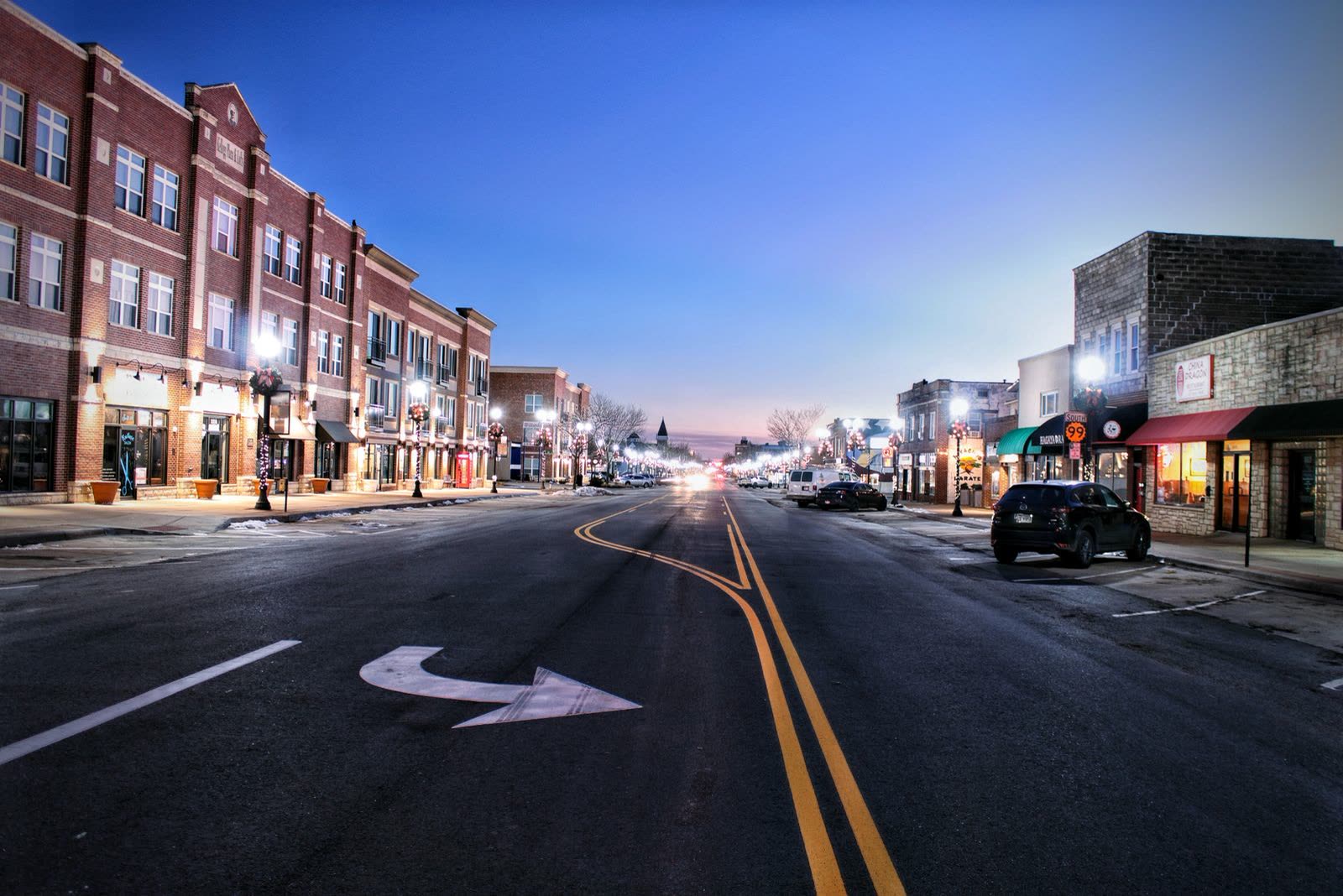 A small-town main street at dusk, shopfronts lit along both sides
